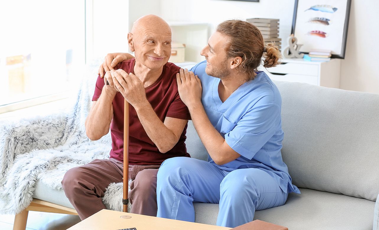 Eine männliche Pflegefachkraft lächelt einen älteren Patienten an, während sie auf einer Couch sitzen