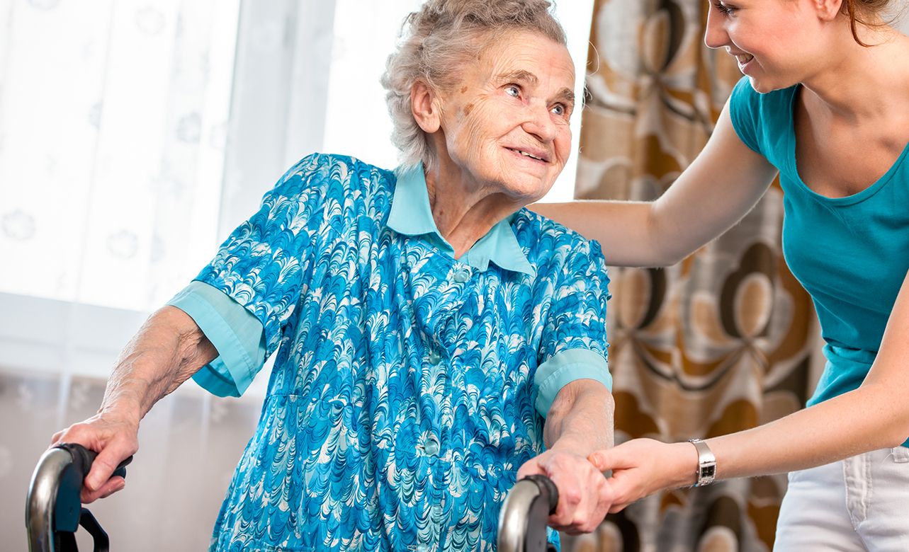Eine Frau unterstützt eine ältere Dame beim Gehen mit einem Rollator